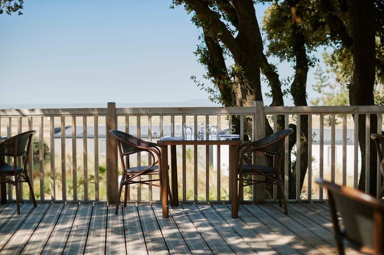 Table en bois avec deux chaises sur une terrasse ensoleillée, entourée d'arbres et offrant une vue dégagée.