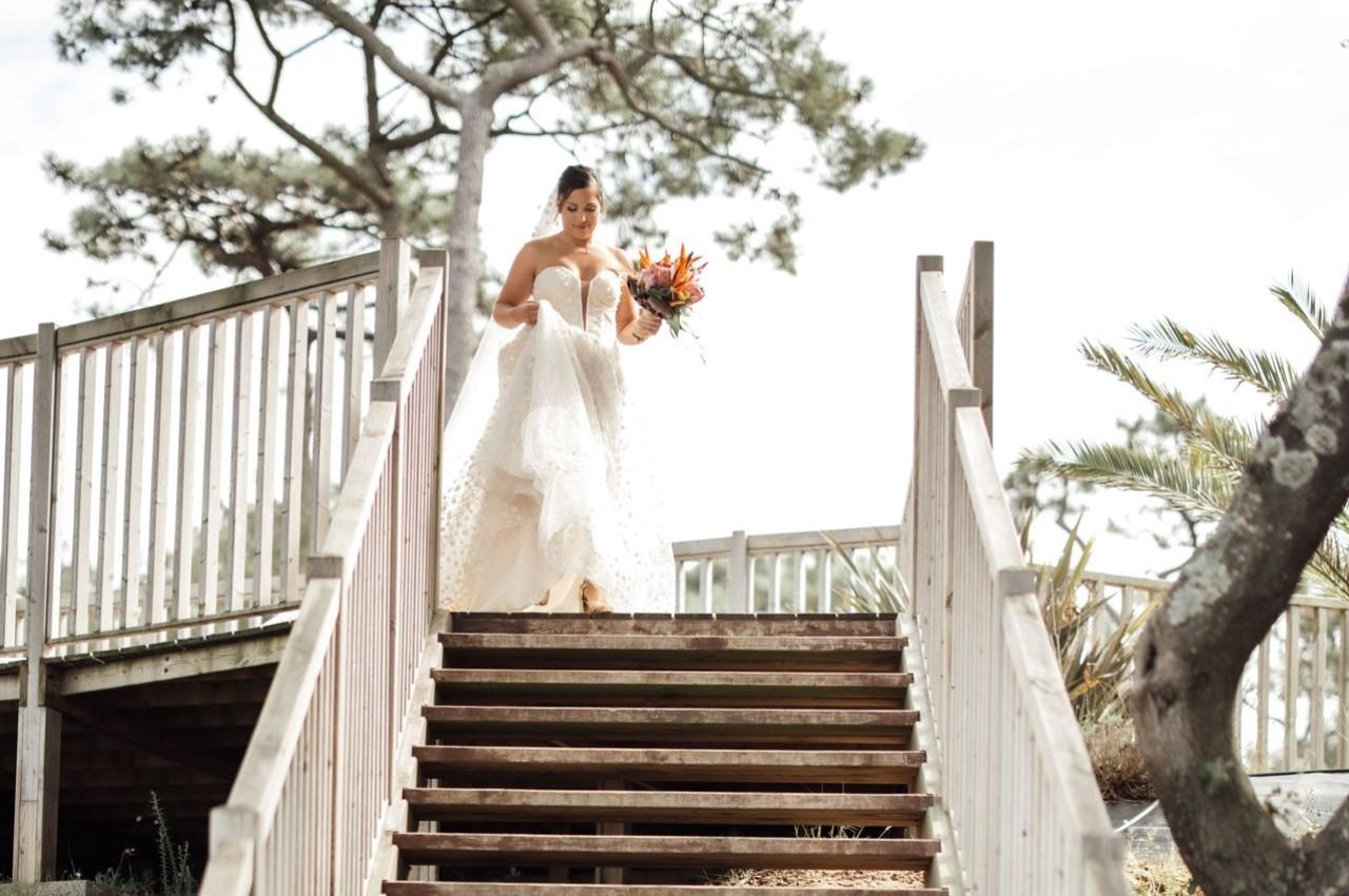 Une mariée en robe blanche descend un escalier en bois, tenant un bouquet coloré, entourée de végétation.