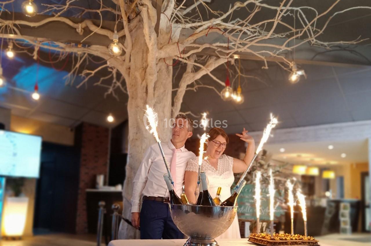 Un couple souriant pose près d'un seau à champagne avec des cierges magiques allumés, sous un arbre décoratif.