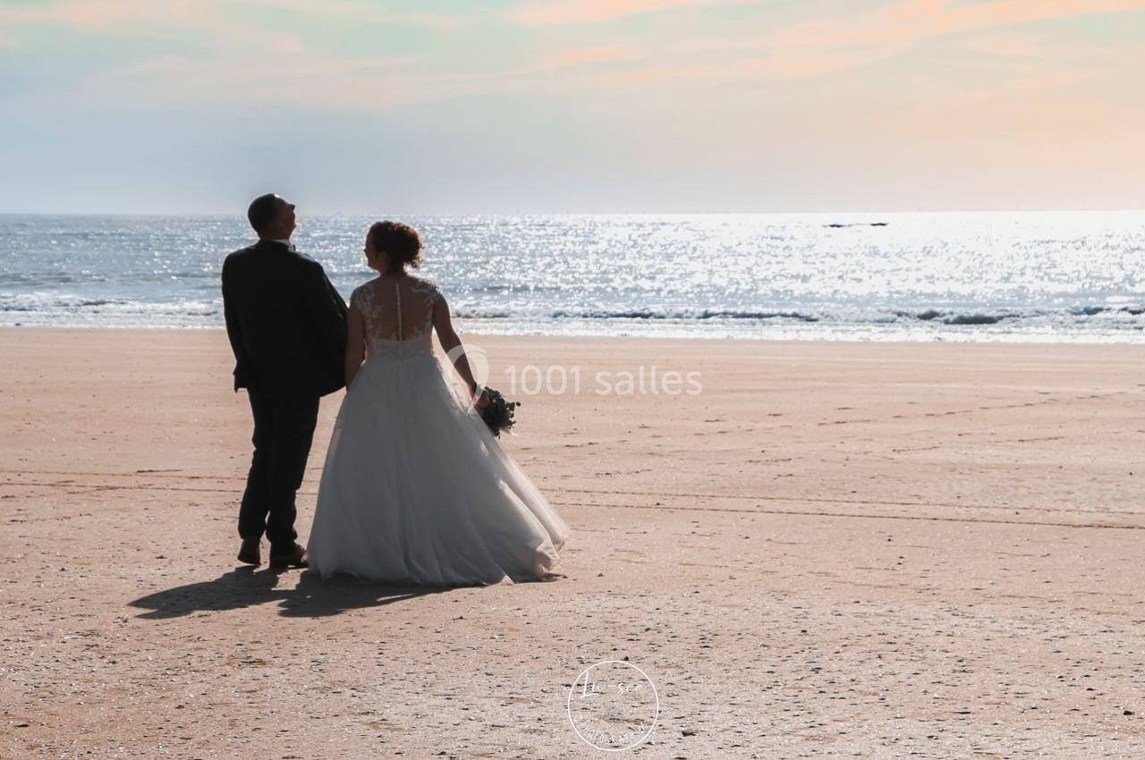 Un couple en tenue de mariage marche sur une plage ensoleillée, face à la mer scintillante.