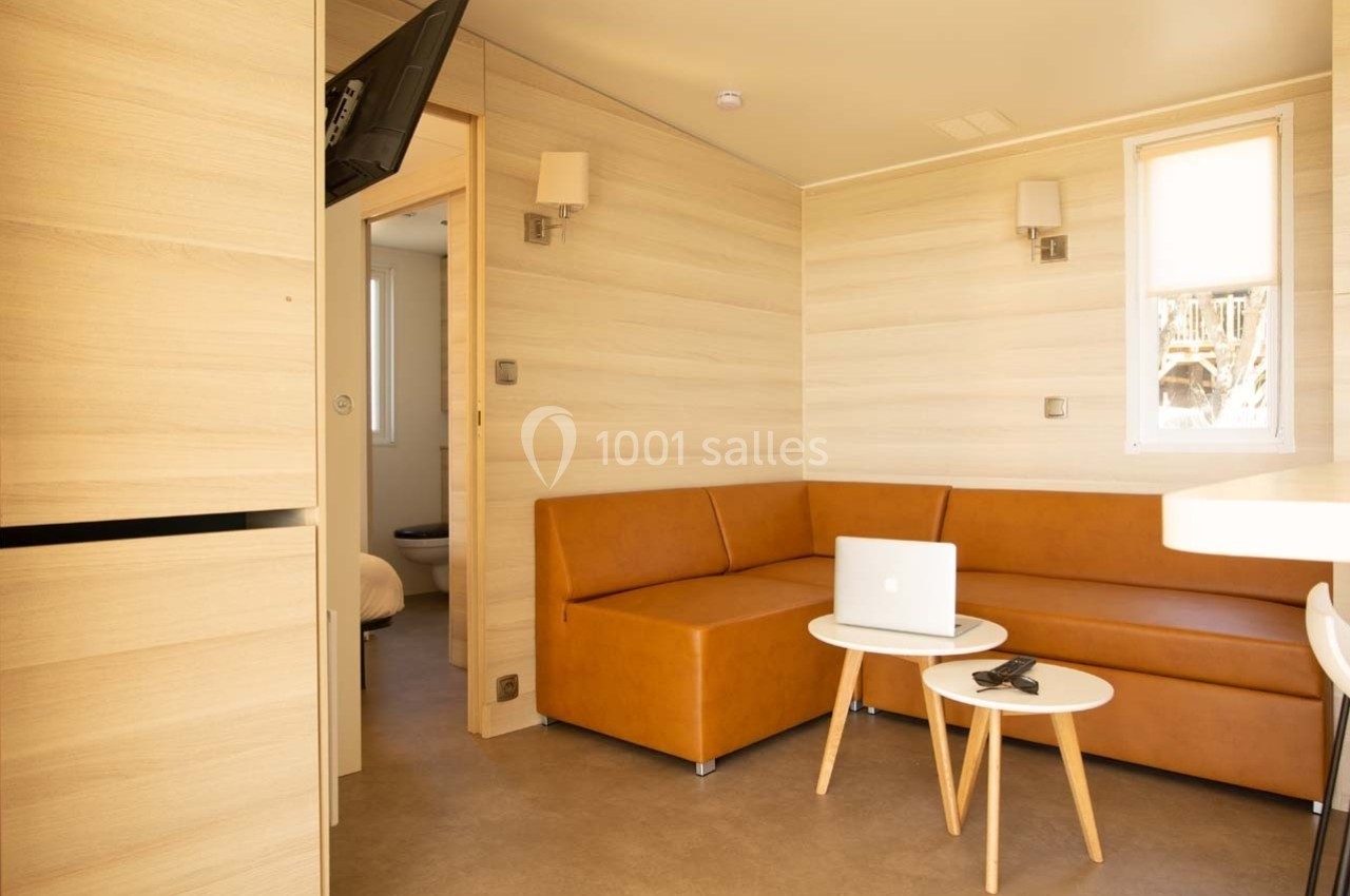 Intérieur lumineux d'un mobil-home avec canapé d'angle, table basse, ordinateur portable et télécommande.