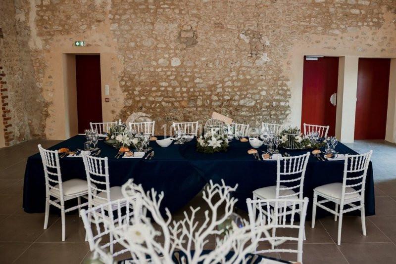 Table dressée avec nappes bleues, chaises blanches et décoration florale, dans une salle aux murs en pierre apparente.