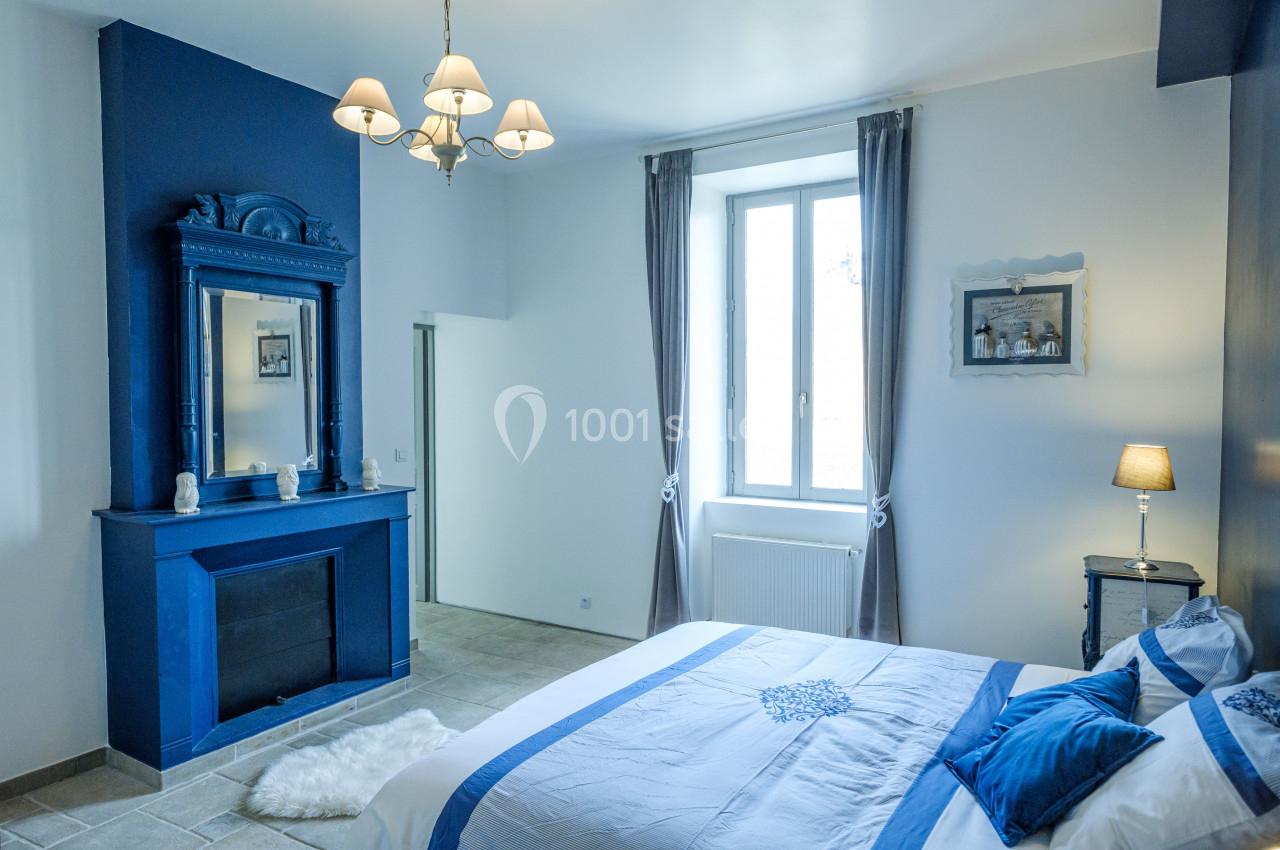 Chambre lumineuse avec murs blancs, cheminée bleue, lit double et grande fenêtre avec rideaux gris.