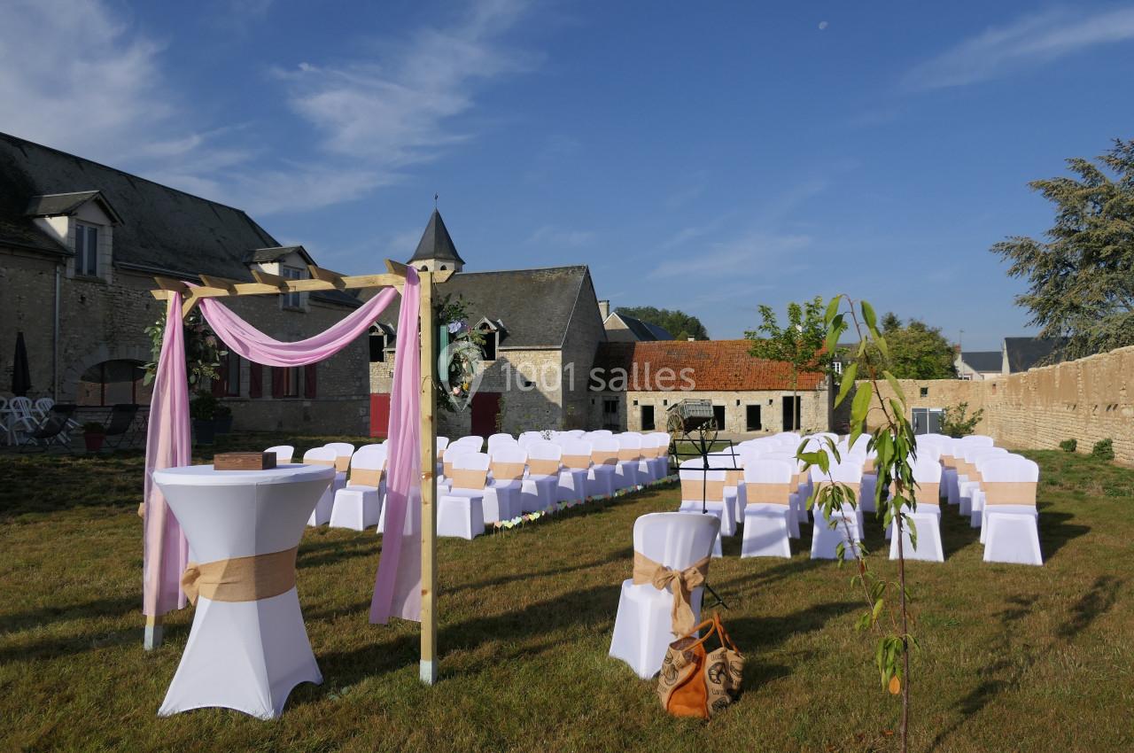 Chaises blanches alignées en extérieur pour une cérémonie, avec arche décorée et bâtiments en pierre en arrière-plan.