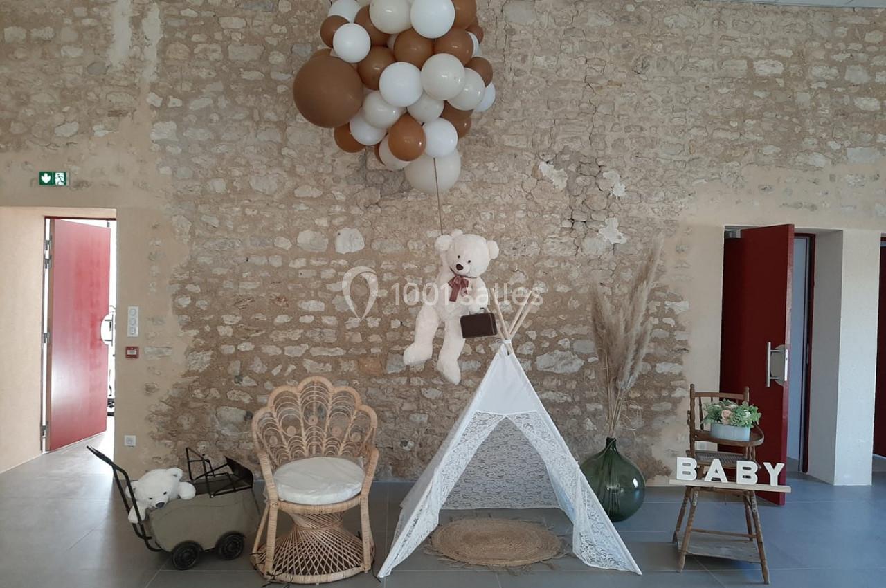 Décoration avec tipi blanc, fauteuil en rotin, ballons suspendus, ours en peluche et lettres formant ’BABY’ sur une table.