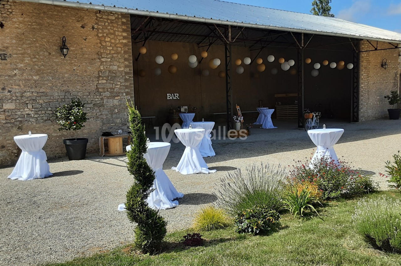 Espace extérieur d'une grange aménagée pour un événement, avec tables hautes drapées de blanc et décorations florales.