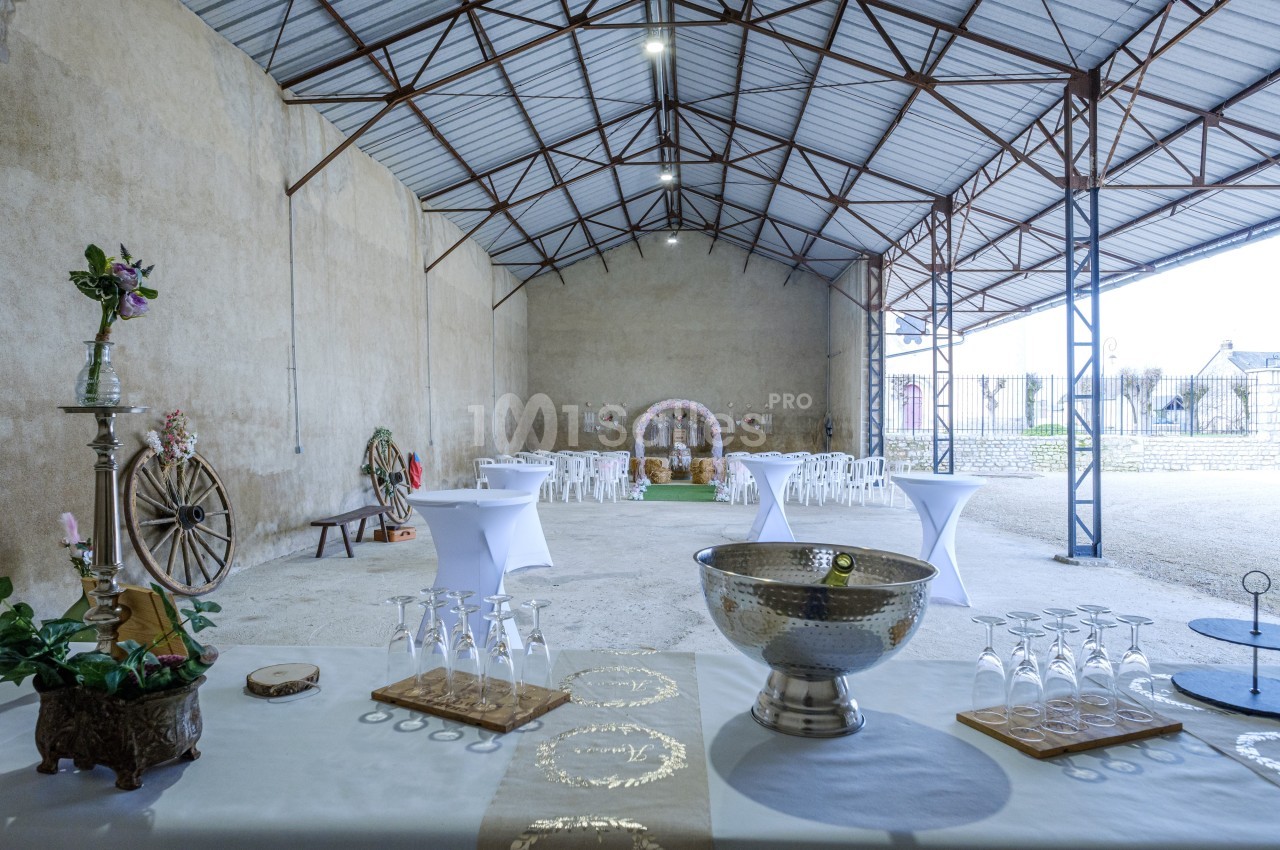 Salle de réception aménagée dans un hangar avec tables dressées, décorations et espace pour un événement festif.