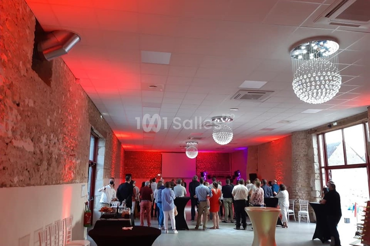 Salle de réception avec des invités debout, éclairée par des lumières rouges et violettes, et des tables hautes disposées.