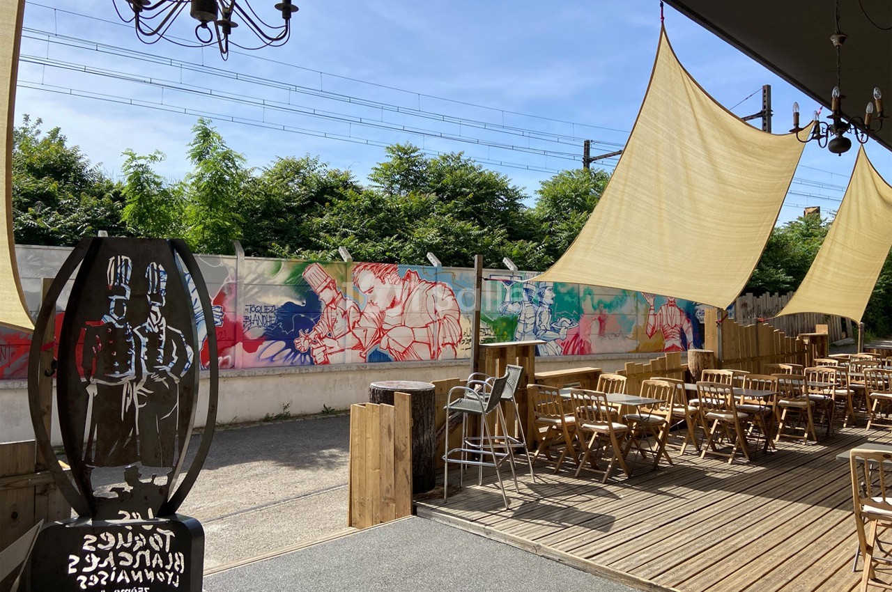 Terrasse en bois avec des tables et chaises, décorée de fresques colorées et ombragée par des voiles tendues.