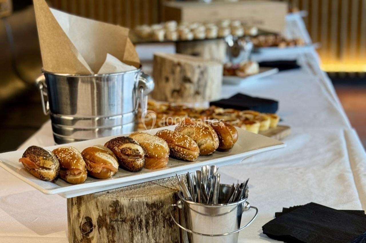 Plateau de bagels garnis sur une table de buffet avec couverts, serviettes et autres plats en arrière-plan.