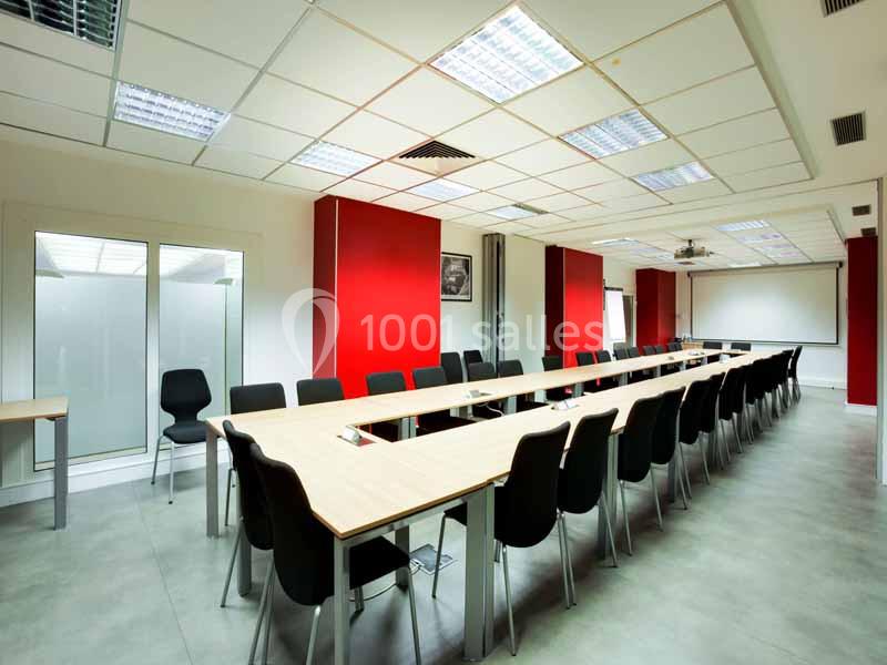 Salle de réunion lumineuse avec une grande table en U, des chaises noires et des murs rouges.