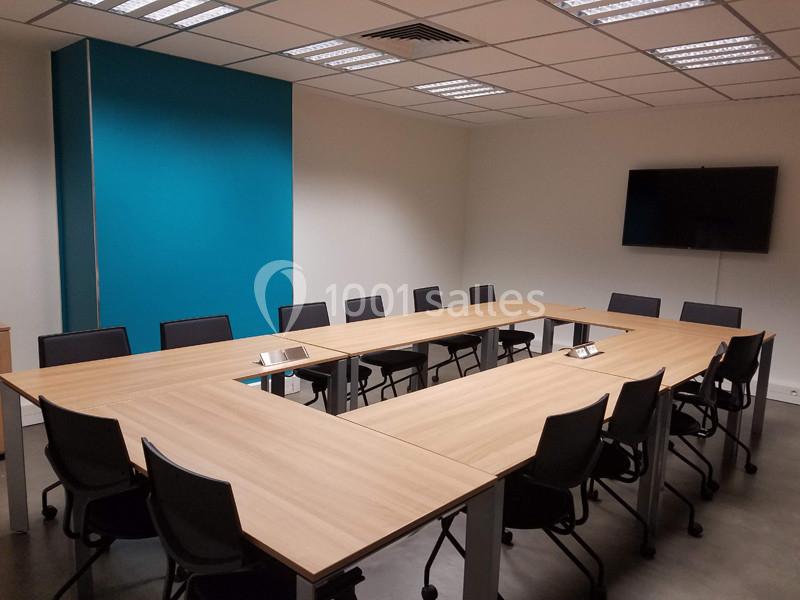 Salle de réunion avec tables en disposition en U, chaises noires, mur bleu et écran mural.