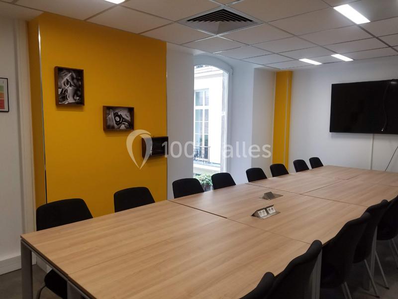Salle de réunion avec une grande table en bois, des chaises noires, un mur jaune et une fenêtre donnant sur l'extérieur.