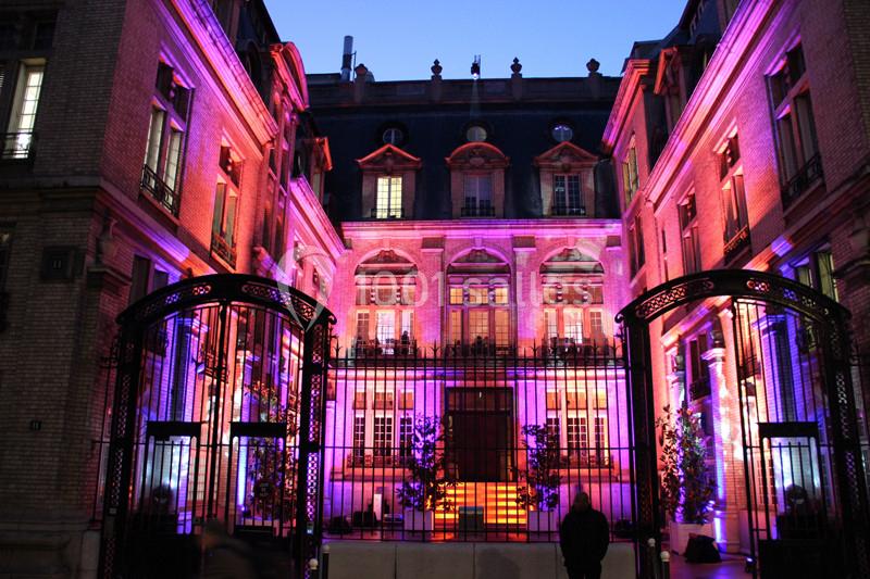 Façade d'un bâtiment historique éclairée par des lumières colorées, vue à travers une grille en fer forgé.