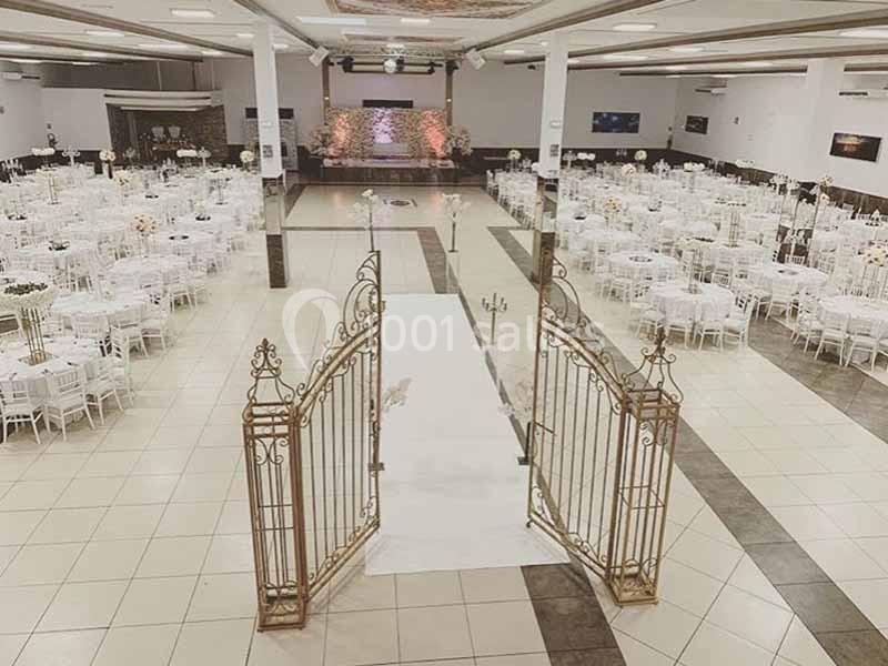 Salle de réception spacieuse avec tables rondes dressées, allée centrale blanche et portail décoratif au premier plan.