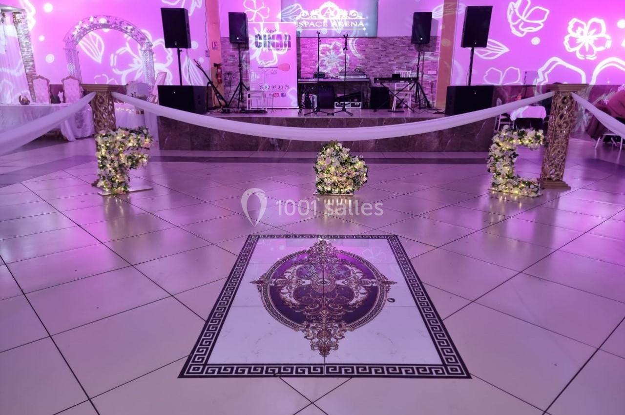 Salle de réception décorée avec des fleurs et éclairages roses, scène équipée de matériel audio en arrière-plan.