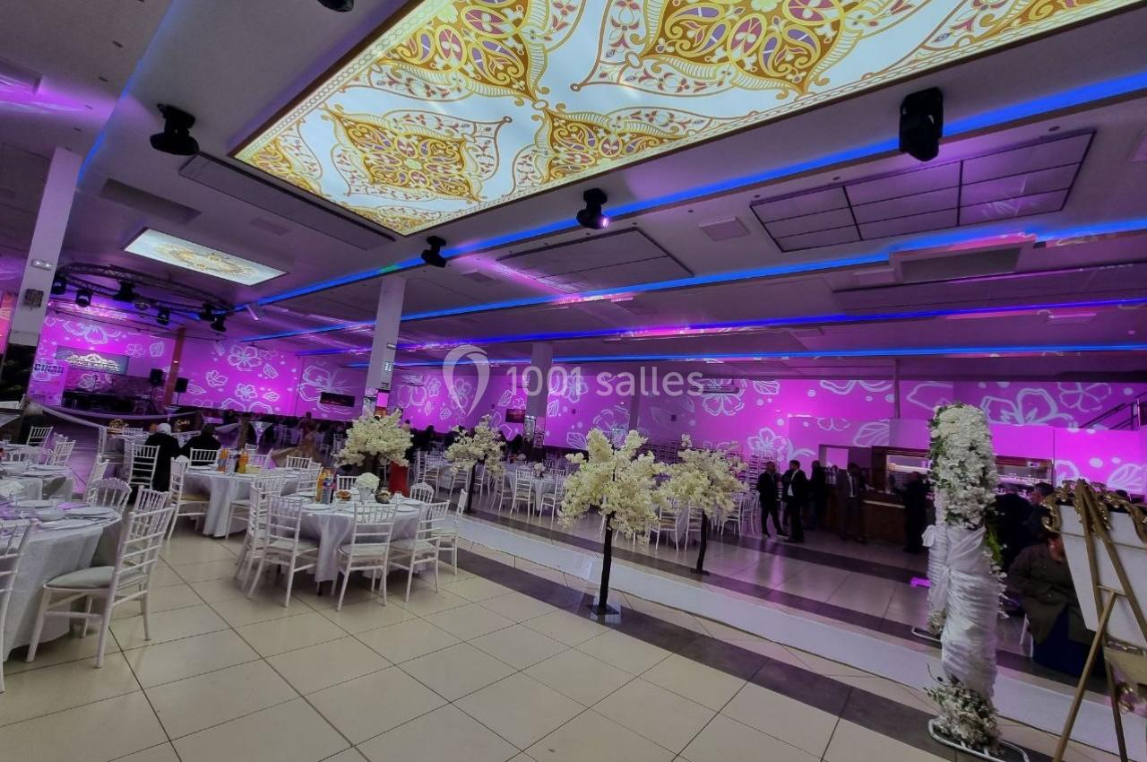 Salle de réception décorée avec des tables dressées, des fleurs blanches et un éclairage rose et violet.