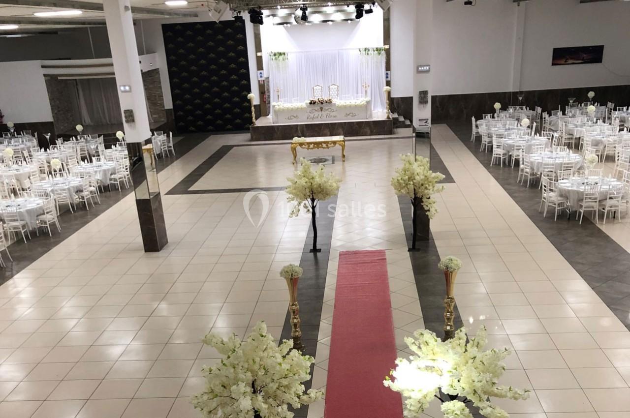 Salle de réception décorée avec des tables blanches, un tapis rouge central et une scène ornée de fleurs blanches.