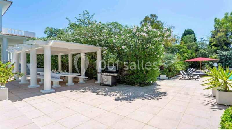 Terrasse extérieure avec pergola blanche, barbecue, chaises longues et végétation luxuriante sous un ciel dégagé.