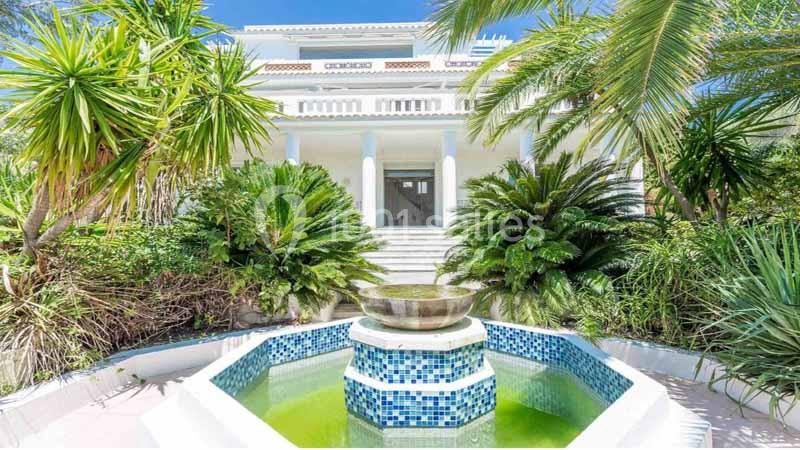 Villa blanche entourée de végétation tropicale avec une fontaine en mosaïque au premier plan.