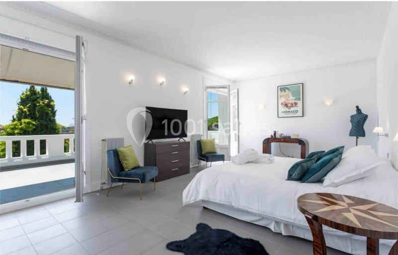 Chambre lumineuse avec lit double, fauteuils, télévision, balcon ouvert sur un jardin et décoration moderne.