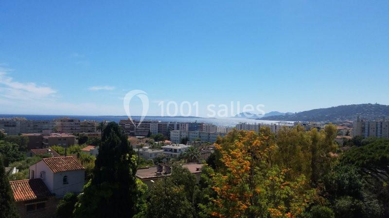 Vue panoramique sur une ville côtière avec des bâtiments, des arbres, la mer et des collines à l'horizon sous un ciel dégagé.