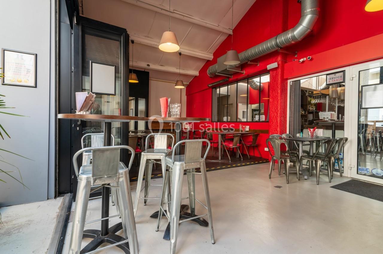 Salle de restaurant moderne avec murs rouges, tables hautes en bois, chaises métalliques et grande baie vitrée.