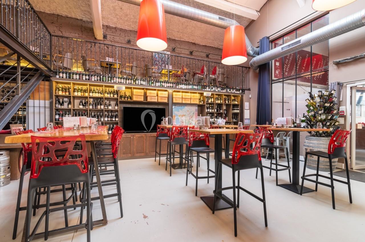 Salle lumineuse avec tables hautes, chaises rouges, étagères remplies de bouteilles de vin et décor industriel.