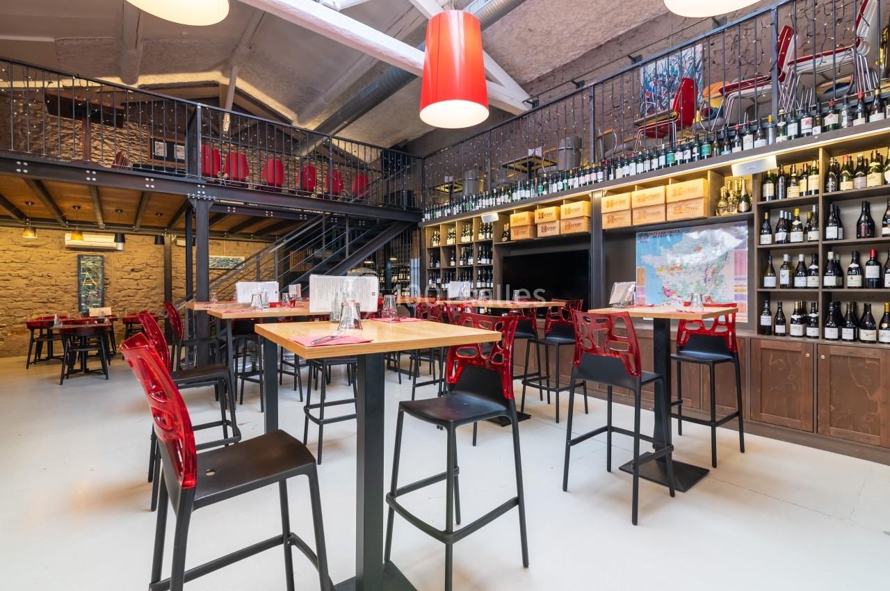 Salle de restaurant moderne avec tables hautes, chaises rouges et noires, étagères remplies de bouteilles de vin.
