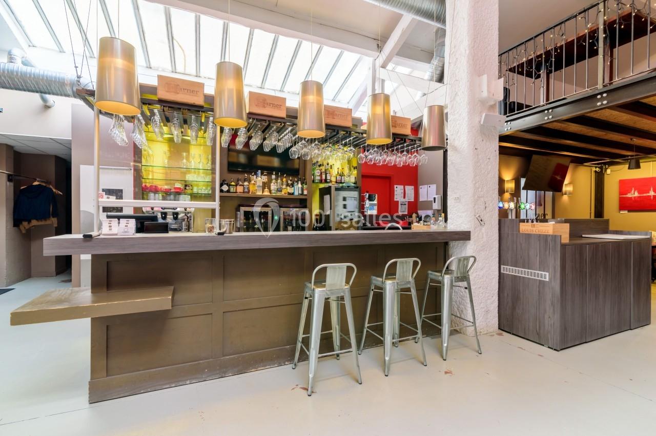Bar moderne avec comptoir en bois, tabourets métalliques, étagères remplies de bouteilles et verres suspendus.
