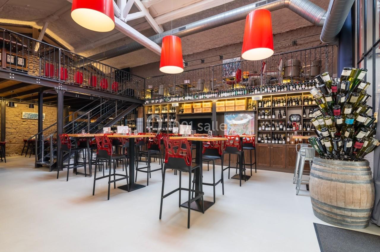 Salle de restaurant moderne avec tables hautes, bar en bois, étagères remplies de bouteilles et luminaires rouges suspendus.