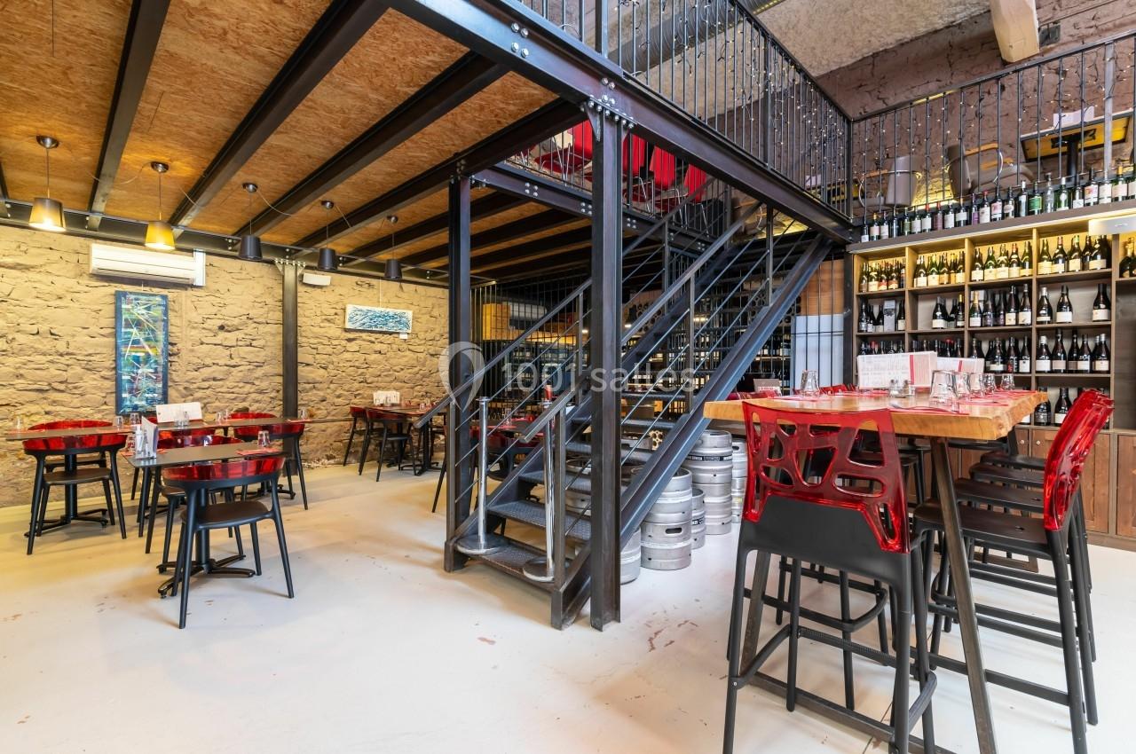 Salle de restaurant moderne avec tables, chaises rouges, étagères de bouteilles et escalier métallique central.