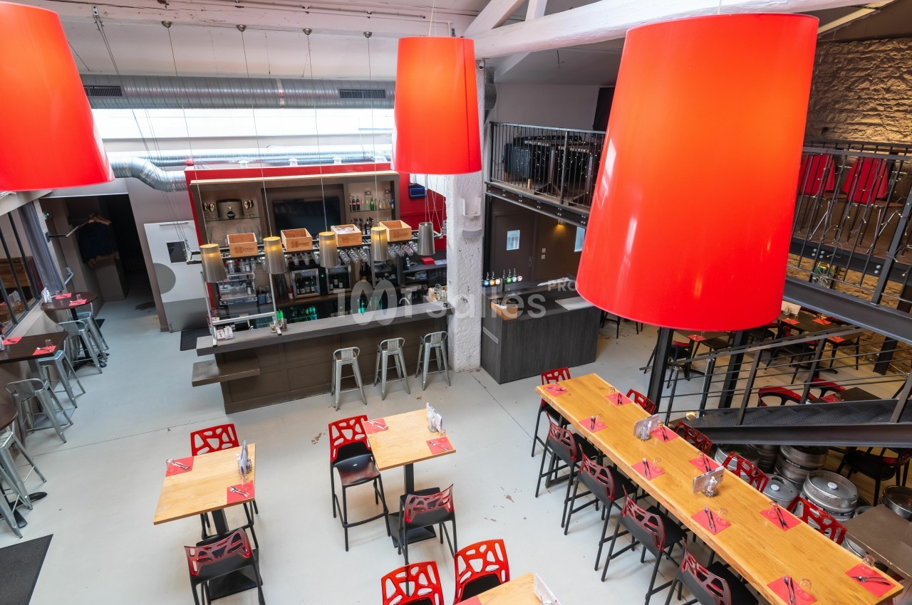 Salle de restaurant moderne avec tables en bois, chaises rouges, bar central et grandes suspensions lumineuses rouges.