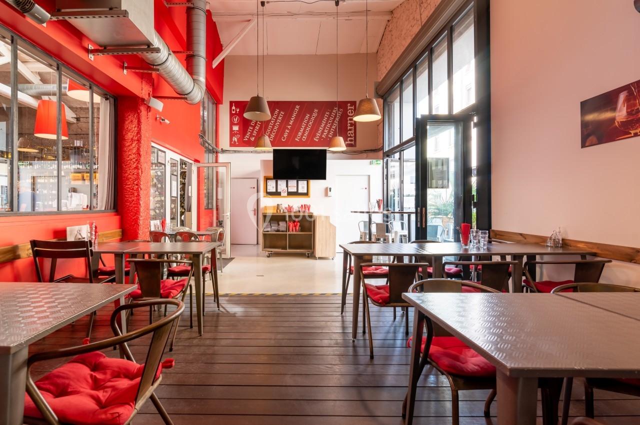 Salle de restaurant lumineuse avec tables en métal, chaises rouges et grandes baies vitrées donnant sur l'extérieur.