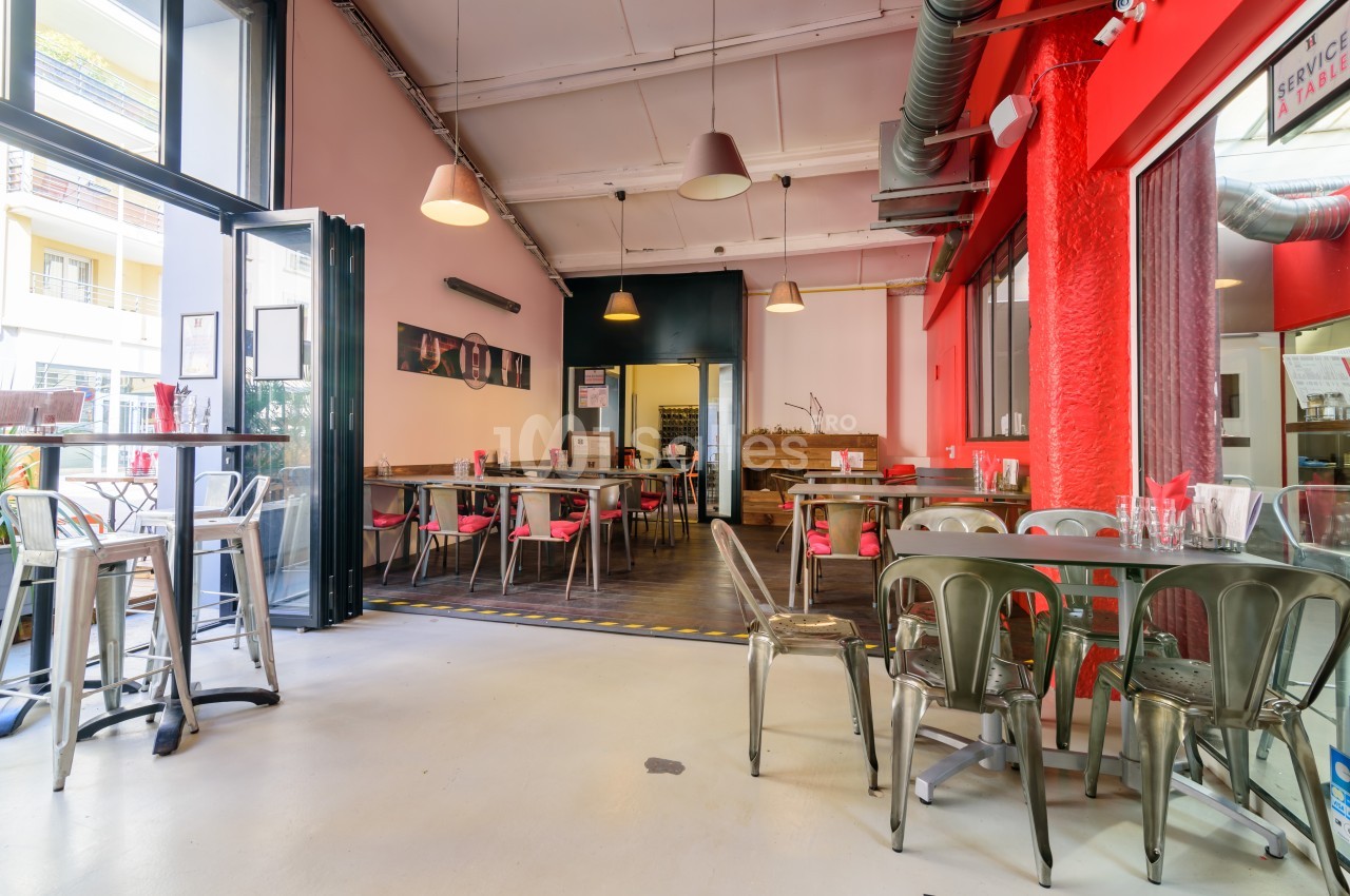 Salle de restaurant lumineuse avec tables en métal et bois, chaises colorées, et murs rouges et roses.