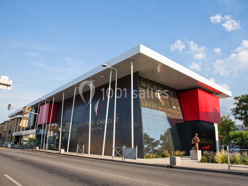 Bâtiment moderne avec façade vitrée et enseigne ’La Cité du Chocolat Valrhona’, situé au bord d'une rue.