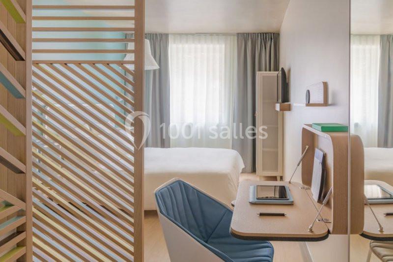 Chambre lumineuse avec bureau, chaise bleue, lit double, cloison ajourée en bois et grande fenêtre avec rideaux.
