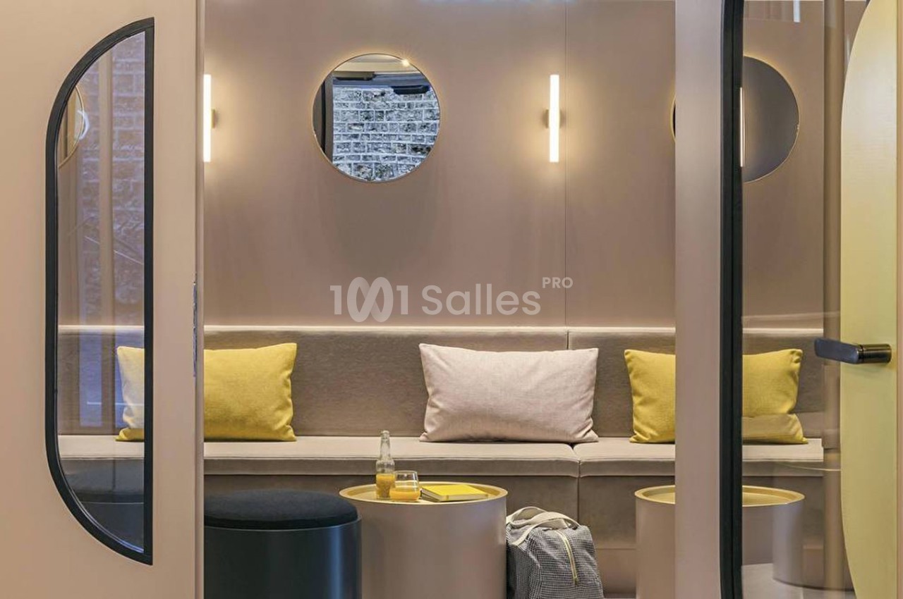 Espace intérieur moderne avec banquette beige, coussins jaunes, tables basses et éclairage mural circulaire.