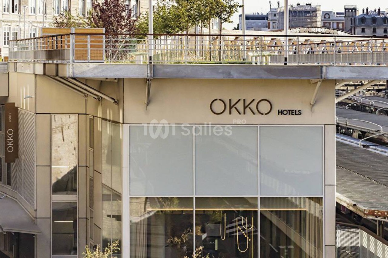 Façade d'un hôtel moderne avec enseigne ’Okko Hotels’, terrasse végétalisée et vue partielle sur des bâtiments urbains.