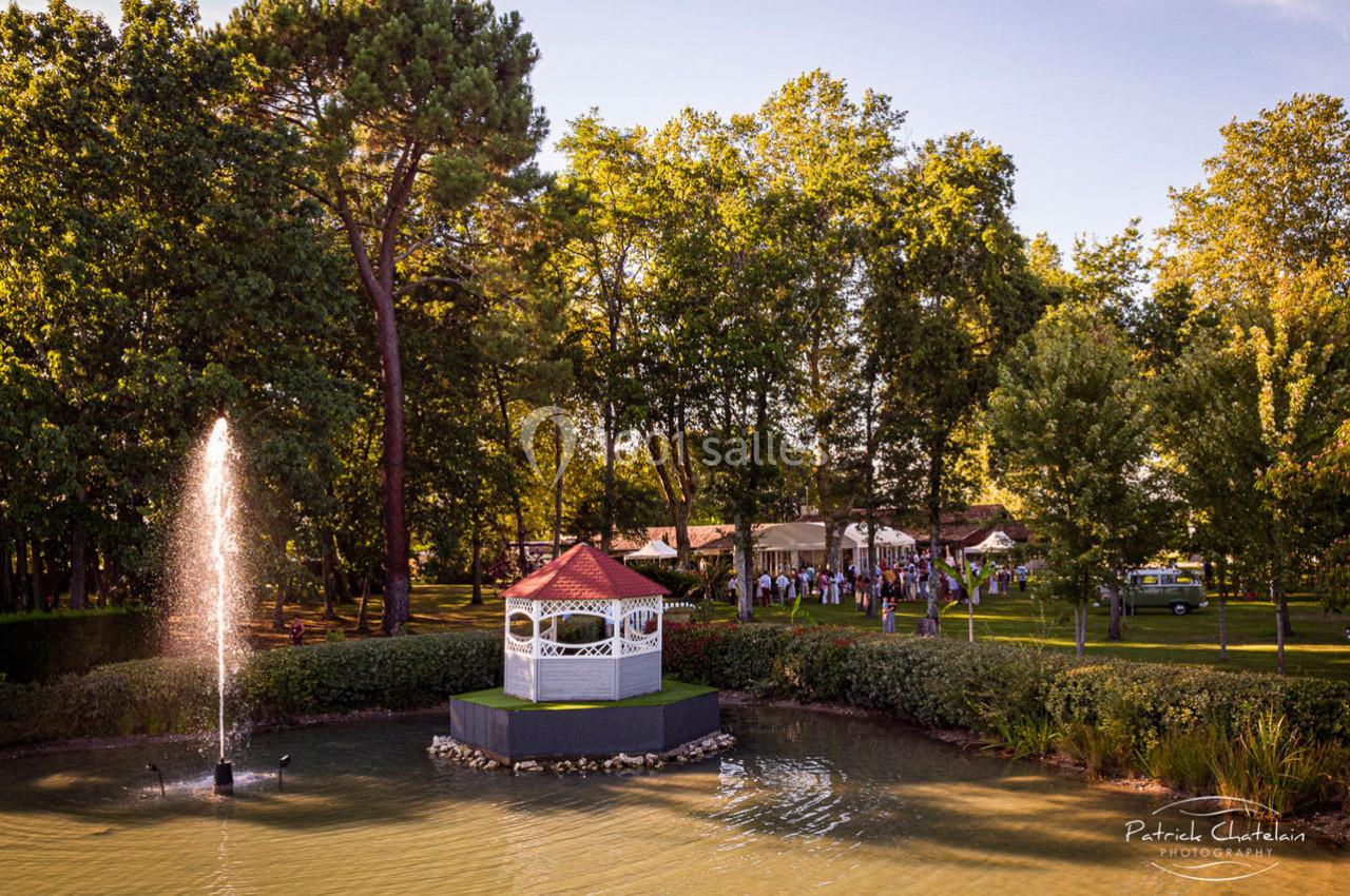 Un kiosque blanc au bord d'un étang avec une fontaine, entouré d'arbres et d'une réception en arrière-plan.