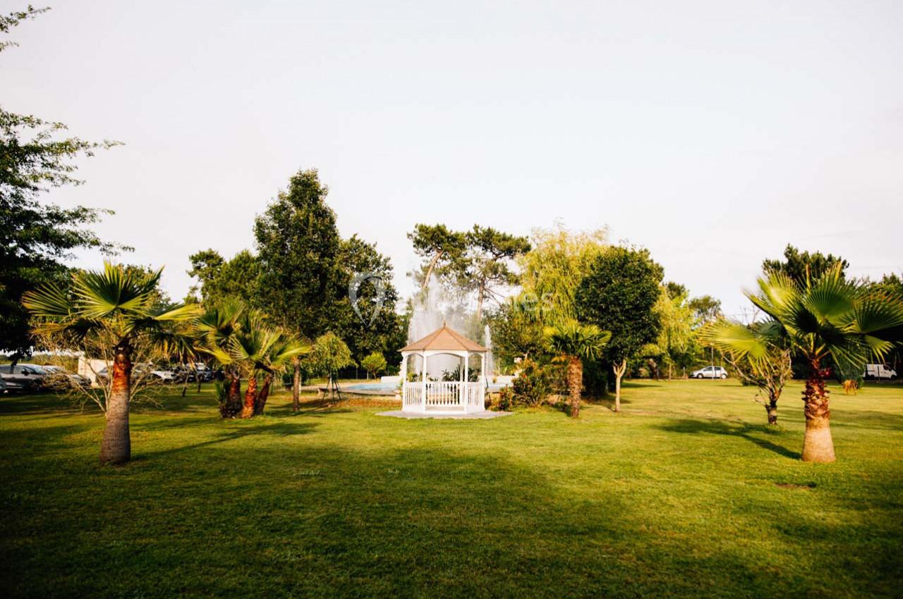 Kiosque blanc entouré de palmiers et d'arbres dans un grand jardin verdoyant sous un ciel clair.