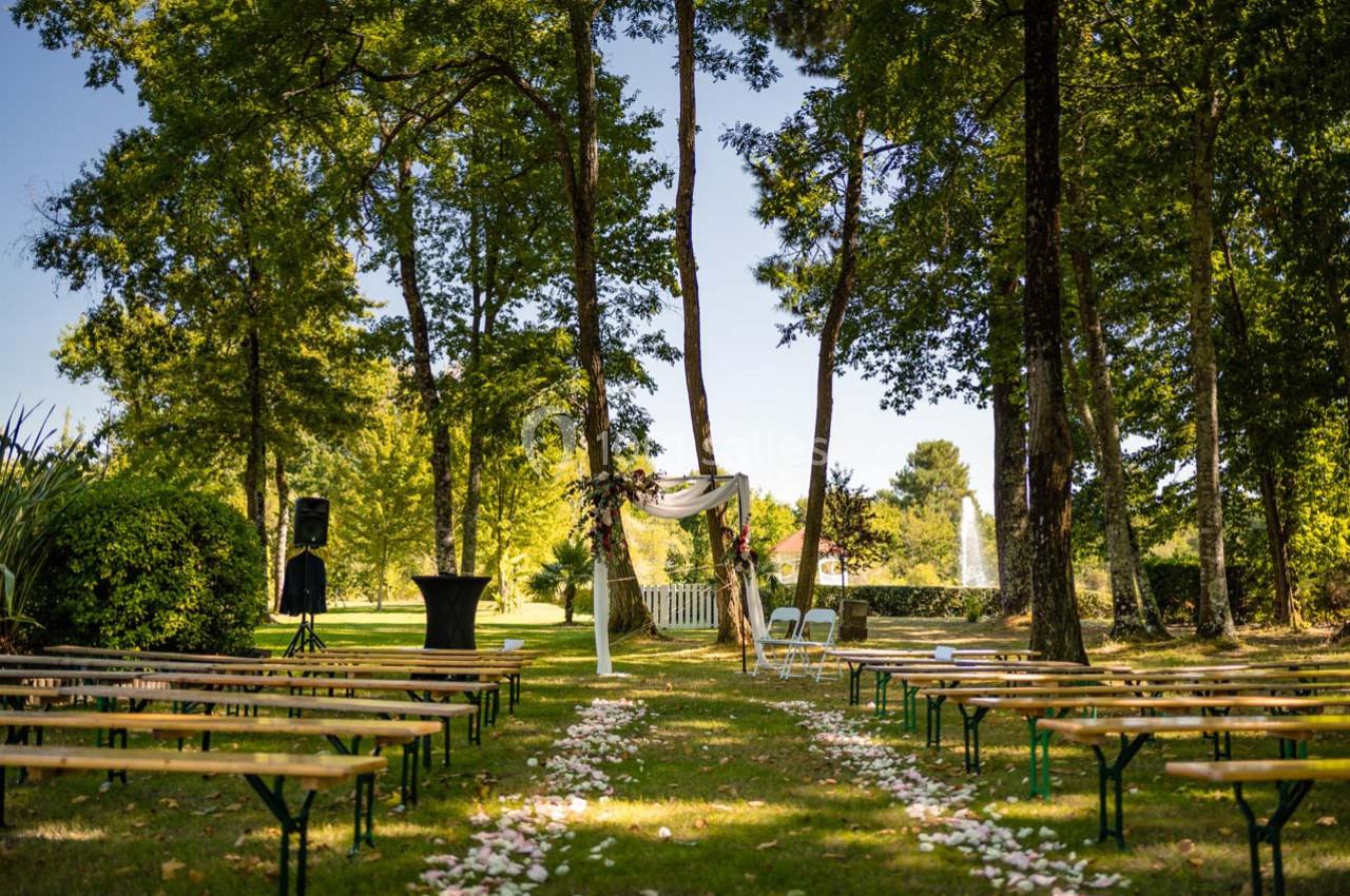 Allée décorée de pétales de fleurs menant à une arche de mariage en extérieur, entourée d'arbres et de bancs en bois.