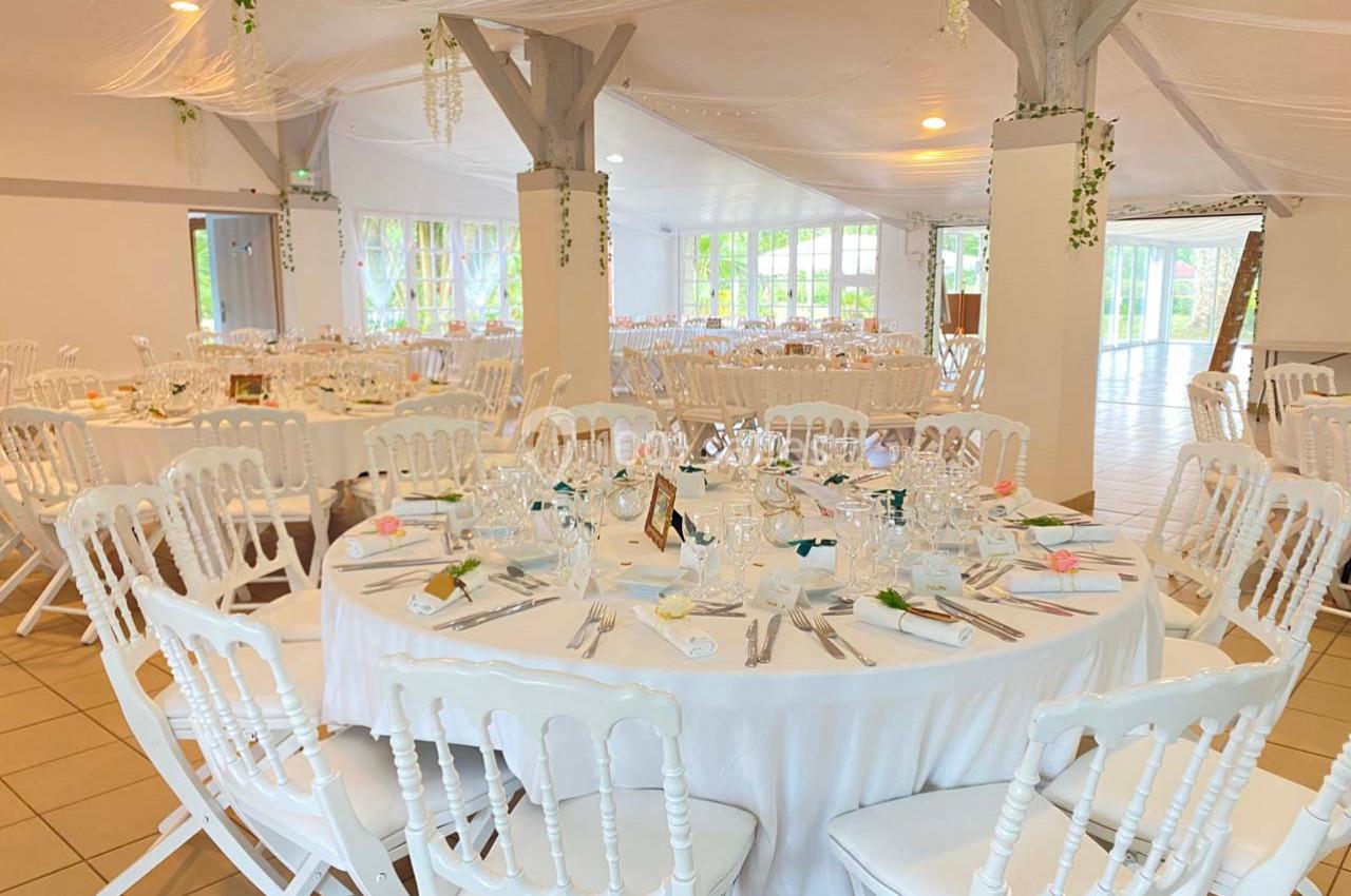 Salle de réception décorée avec des tables rondes dressées, nappes blanches et chaises blanches, ambiance lumineuse.