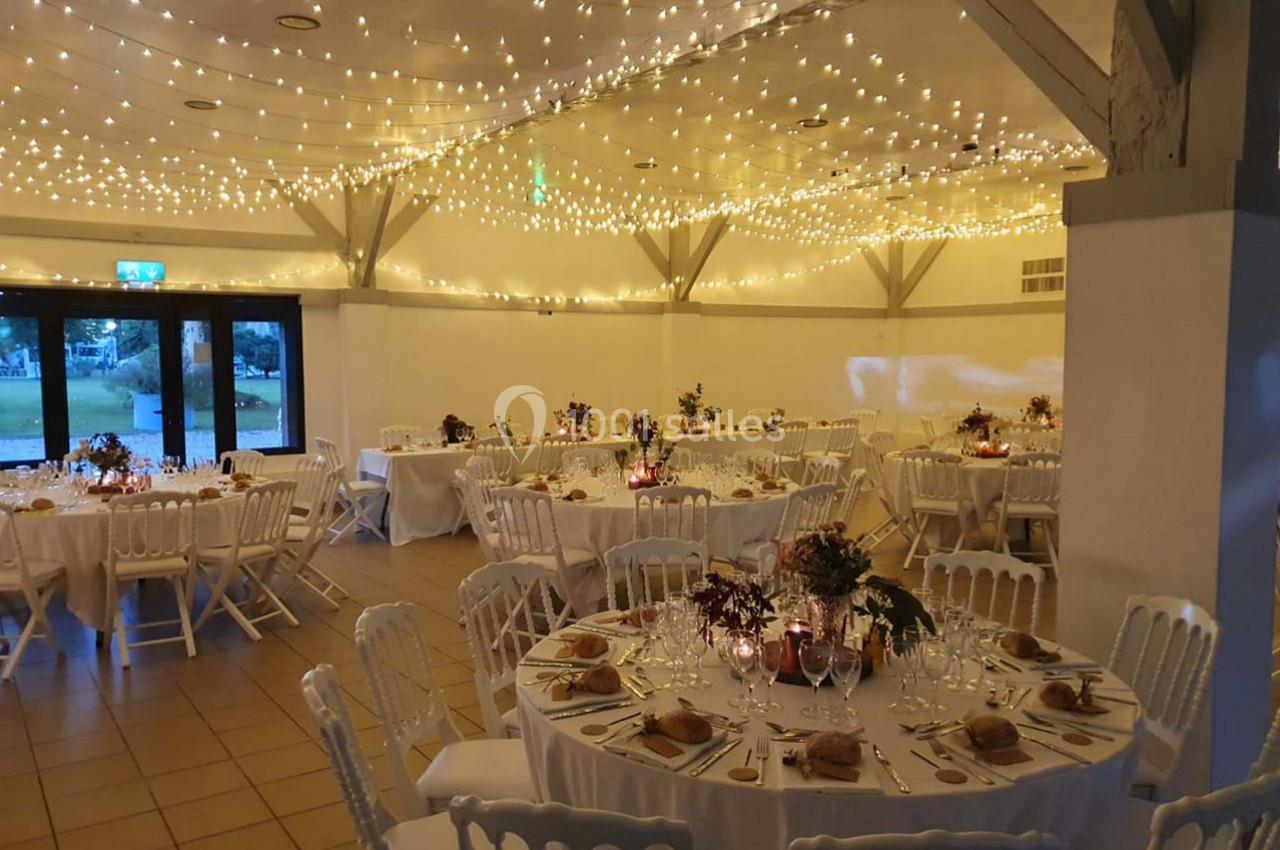 Salle de réception décorée pour un mariage, avec tables rondes dressées et guirlandes lumineuses suspendues au plafond.