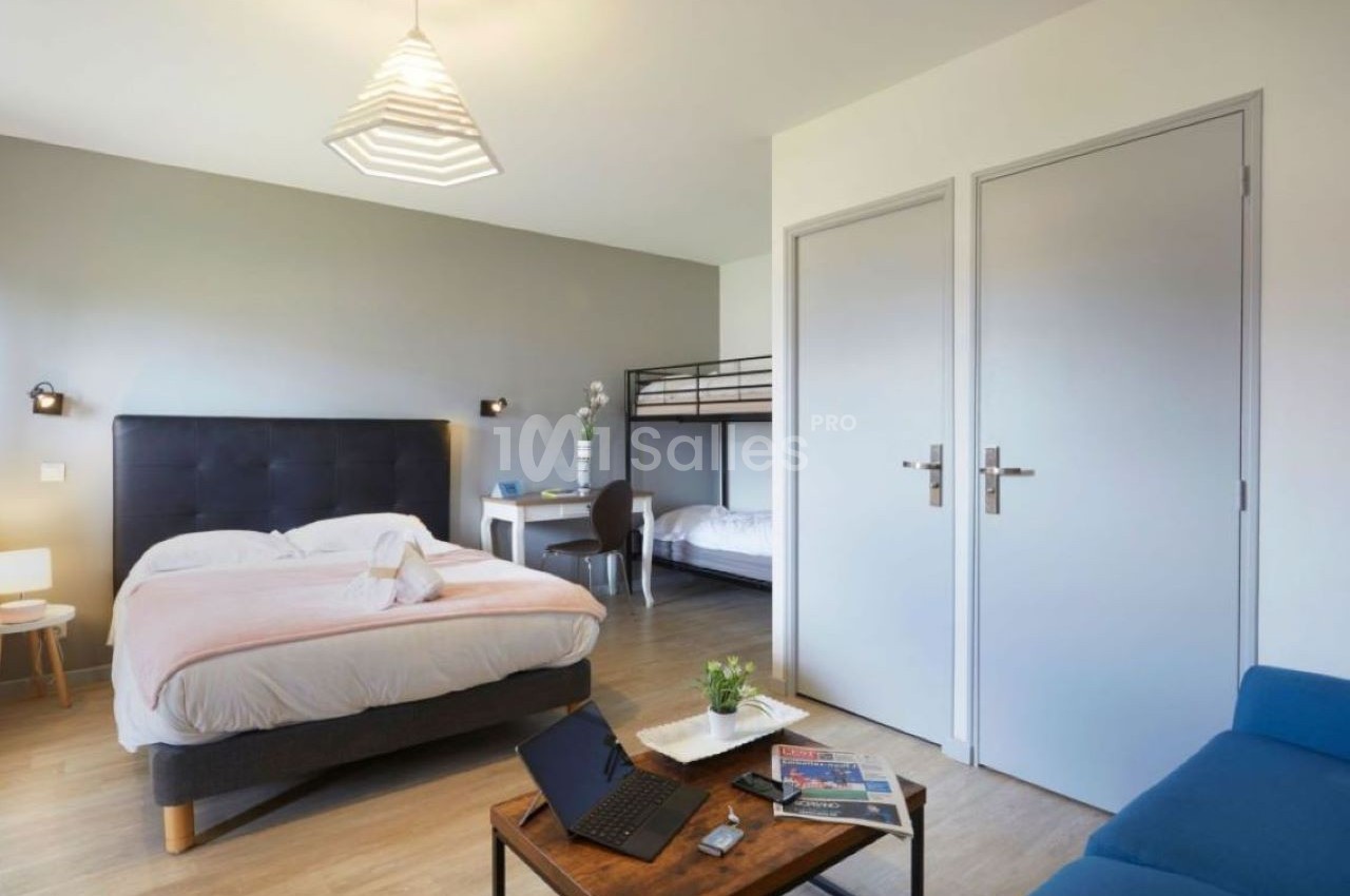 Chambre lumineuse avec lit double, lits superposés, bureau, canapé bleu et table basse avec ordinateur portable.