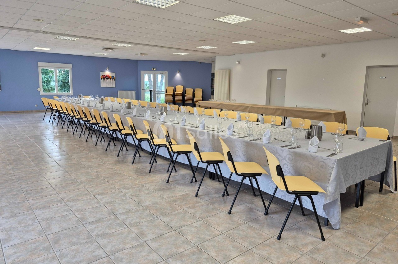 Grande salle avec deux longues tables dressées pour un repas, chaises jaunes et murs clairs.