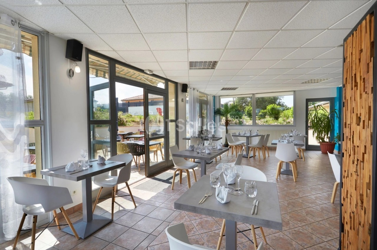 Salle de restaurant lumineuse avec tables dressées, grandes baies vitrées et vue sur un espace extérieur verdoyant.