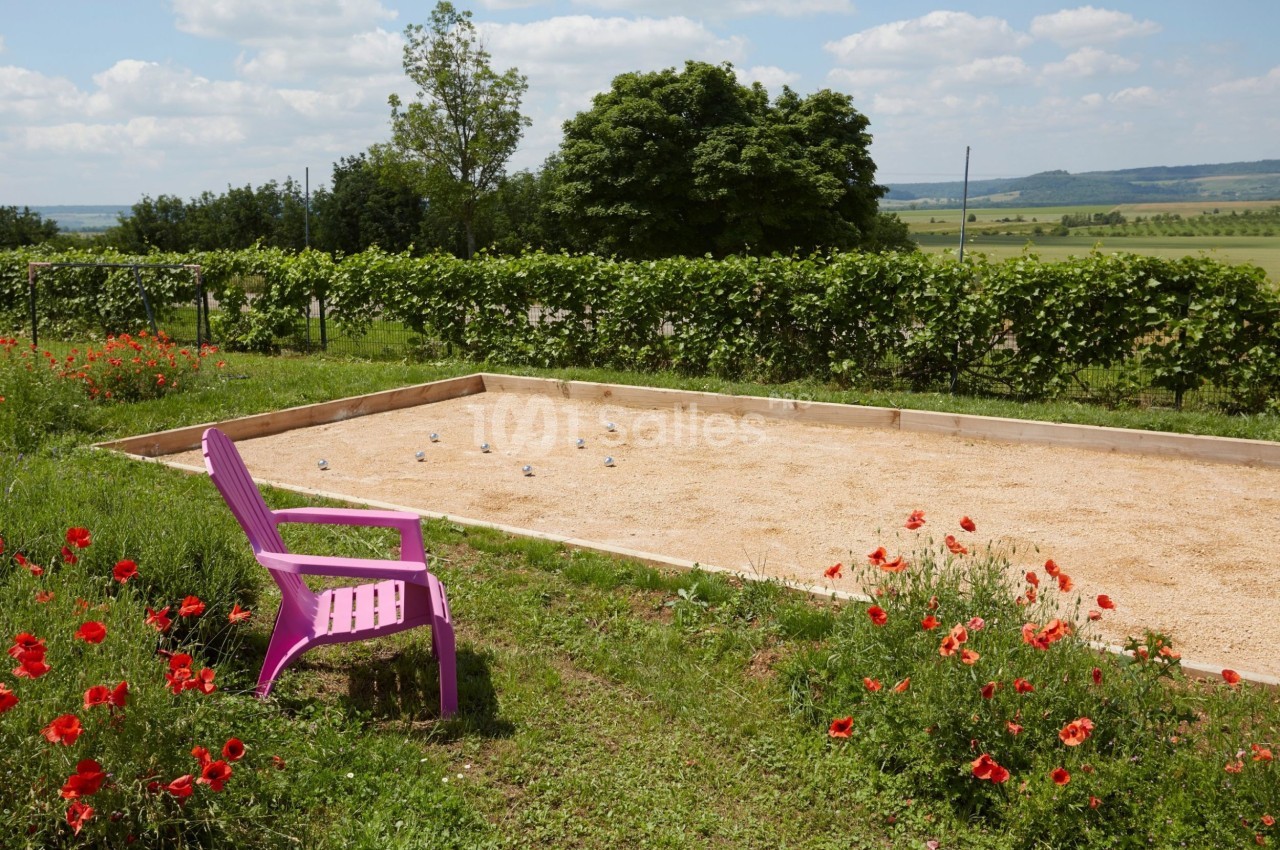 Terrain de pétanque en gravier entouré de verdure et de coquelicots, avec une chaise rose au premier plan.