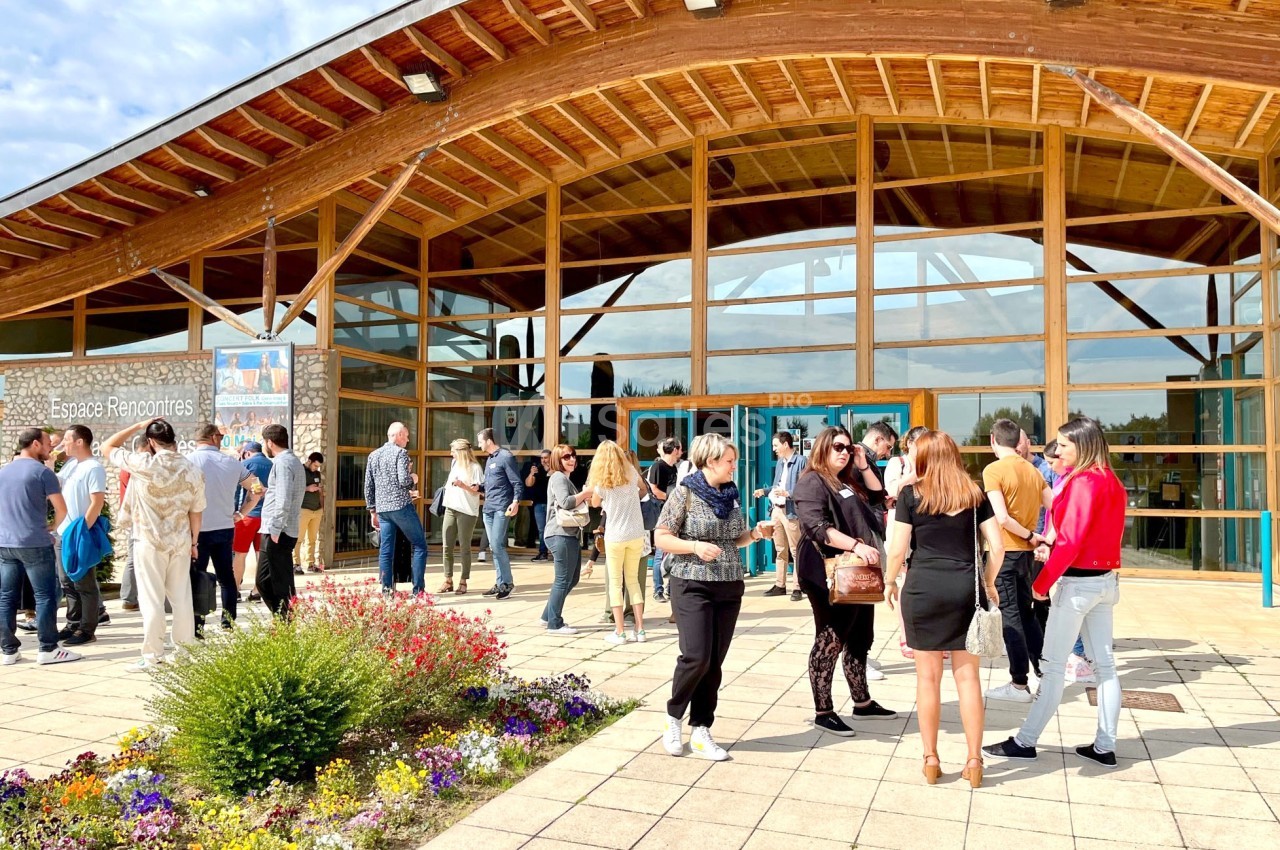 Groupe de personnes discutant devant un bâtiment en bois avec une façade vitrée, entouré de fleurs et d'une terrasse pavée.