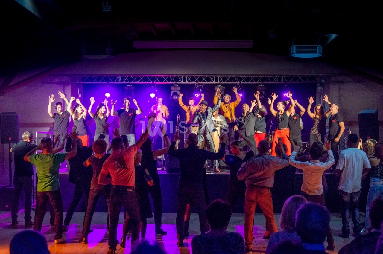 Groupe de musiciens et chanteurs sur scène avec un public debout levant les bras dans une ambiance festive en intérieur.